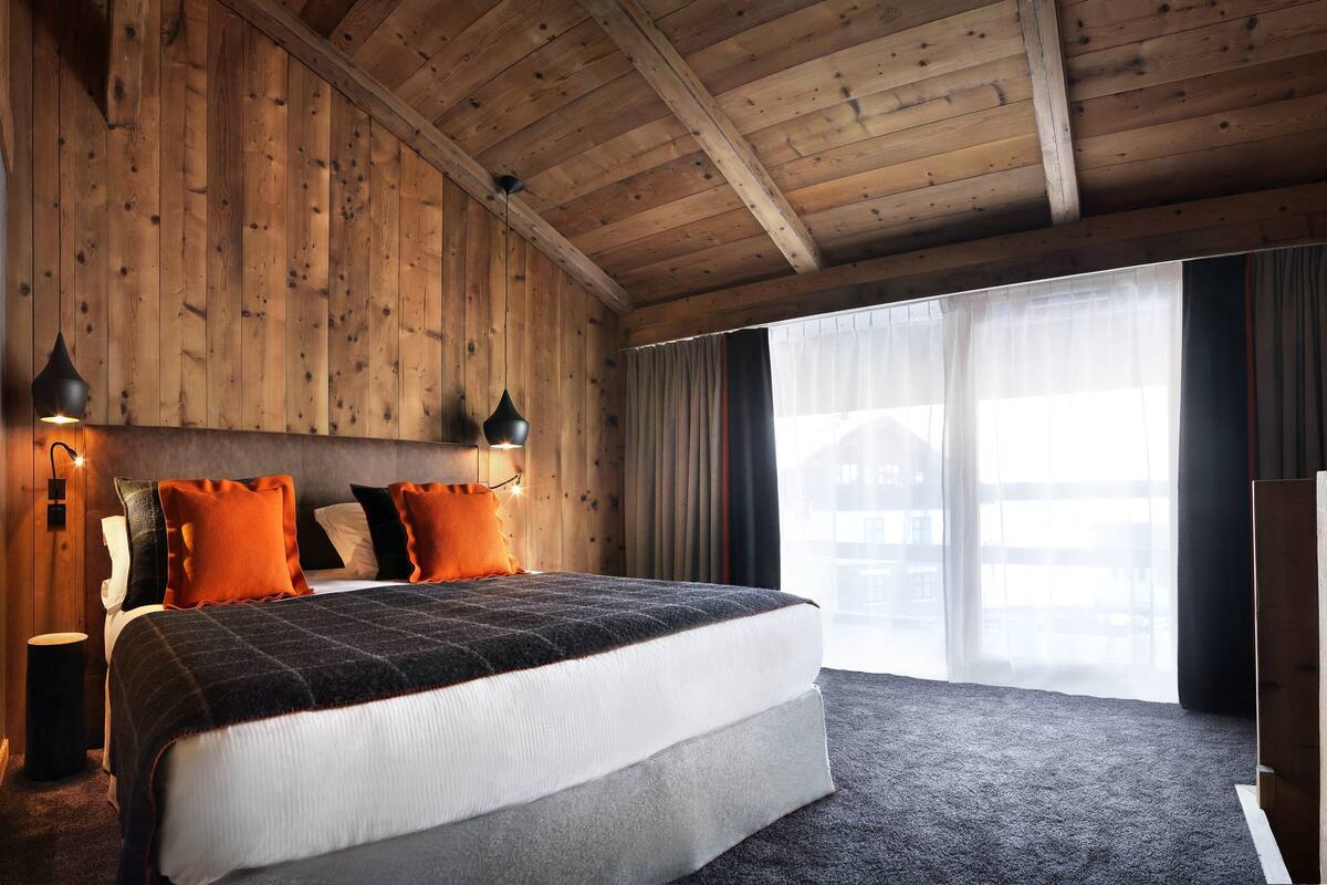 A wooden-paneled bedroom with a large bed, two bedside lamps, and a view of a building through a window.