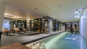 Un espace piscine intérieur moderne, avec des murs en pierre, des portes vitrées et des chaises longues en bois.