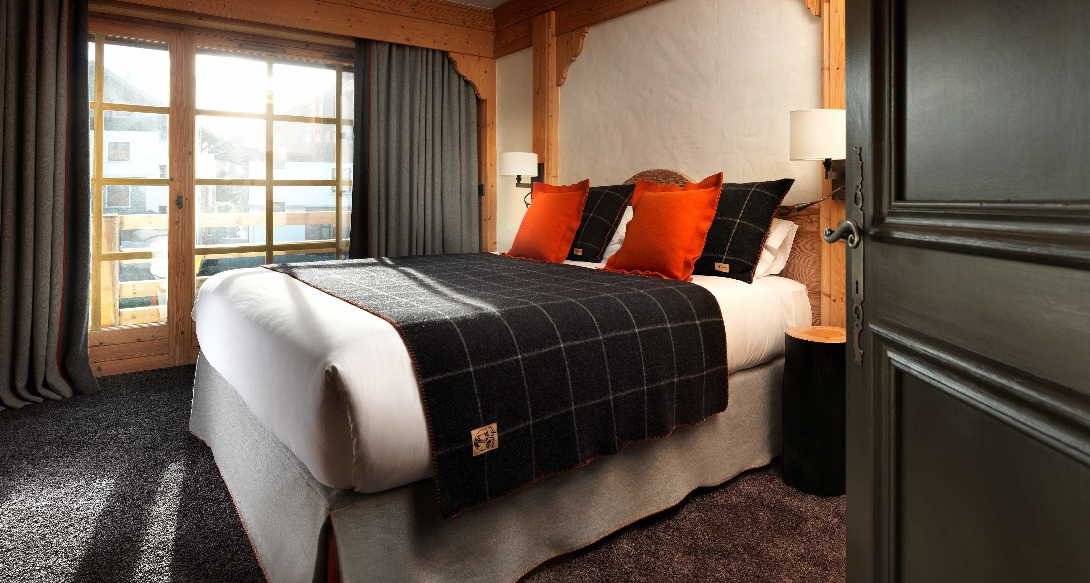 A hotel room with a large bed, a wooden headboard, a nightstand with a lamp, and a view through a window with curtains.