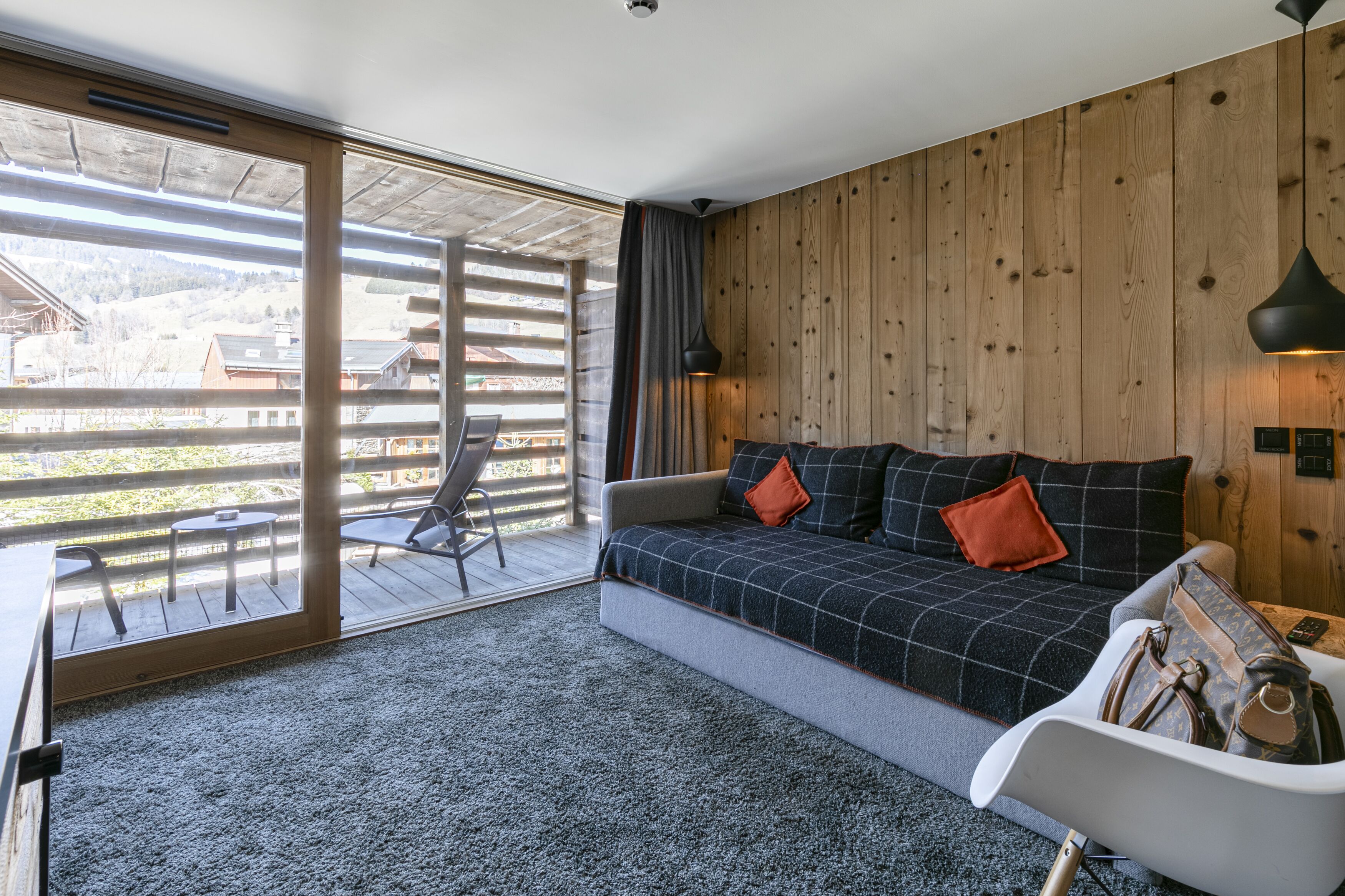 Um quarto aconchegante com sofá, uma mesinha e uma cadeira. Há uma janela grande com vista para o exterior.