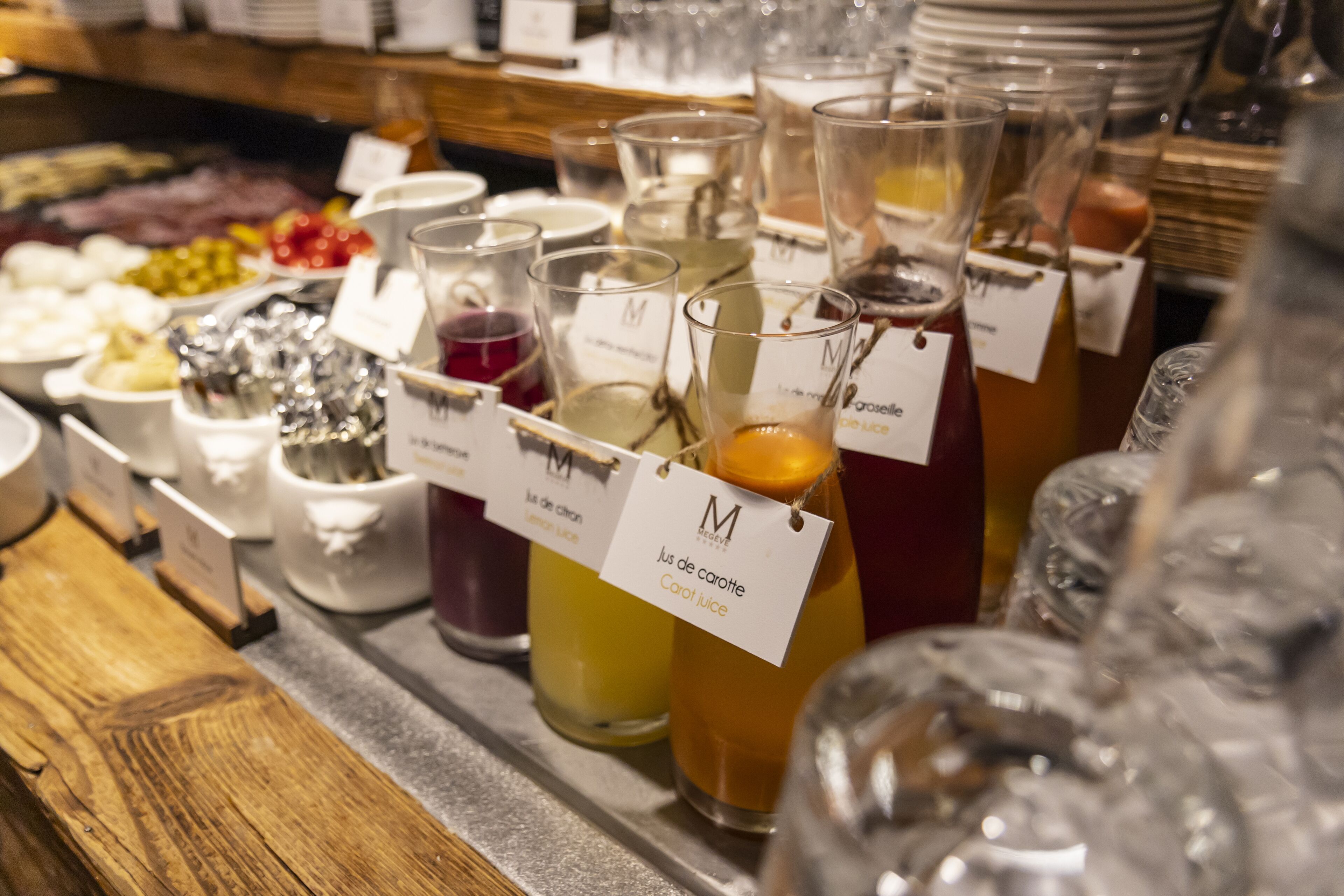 A variety of beverages in glass containers with labels, a buffet area with fruits and cheeses, and a wooden counter.