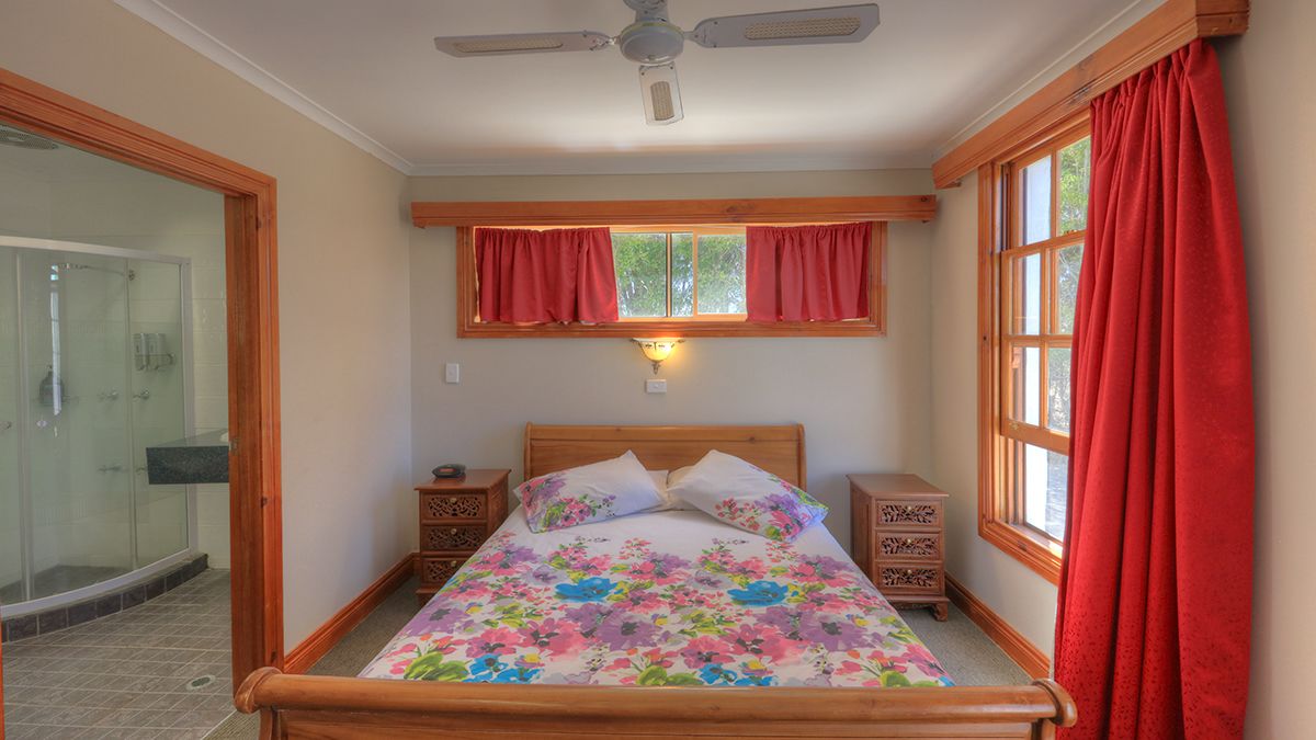 Un dormitorio con cama de madera, ventana con cortinas rojas, ventilador de techo y mesita de noche con lámpara.