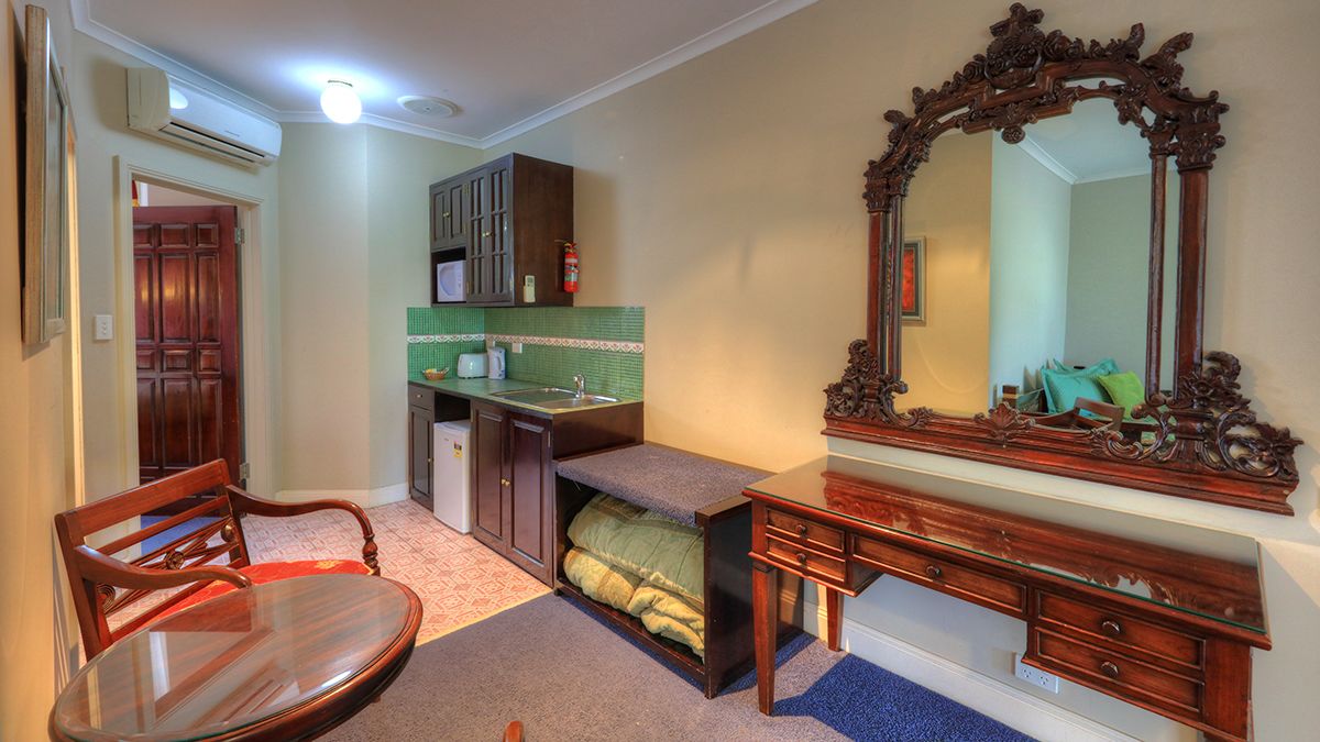 A compact bedroom with a wooden bed, a small desk with drawers, a large ornate mirror, and a built-in bench with storage.