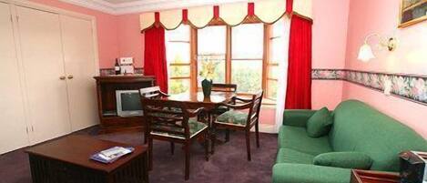 A living room with a green sofa, a wooden coffee table, a dining table with chairs, a TV on a cabinet, and a window with red curtains.