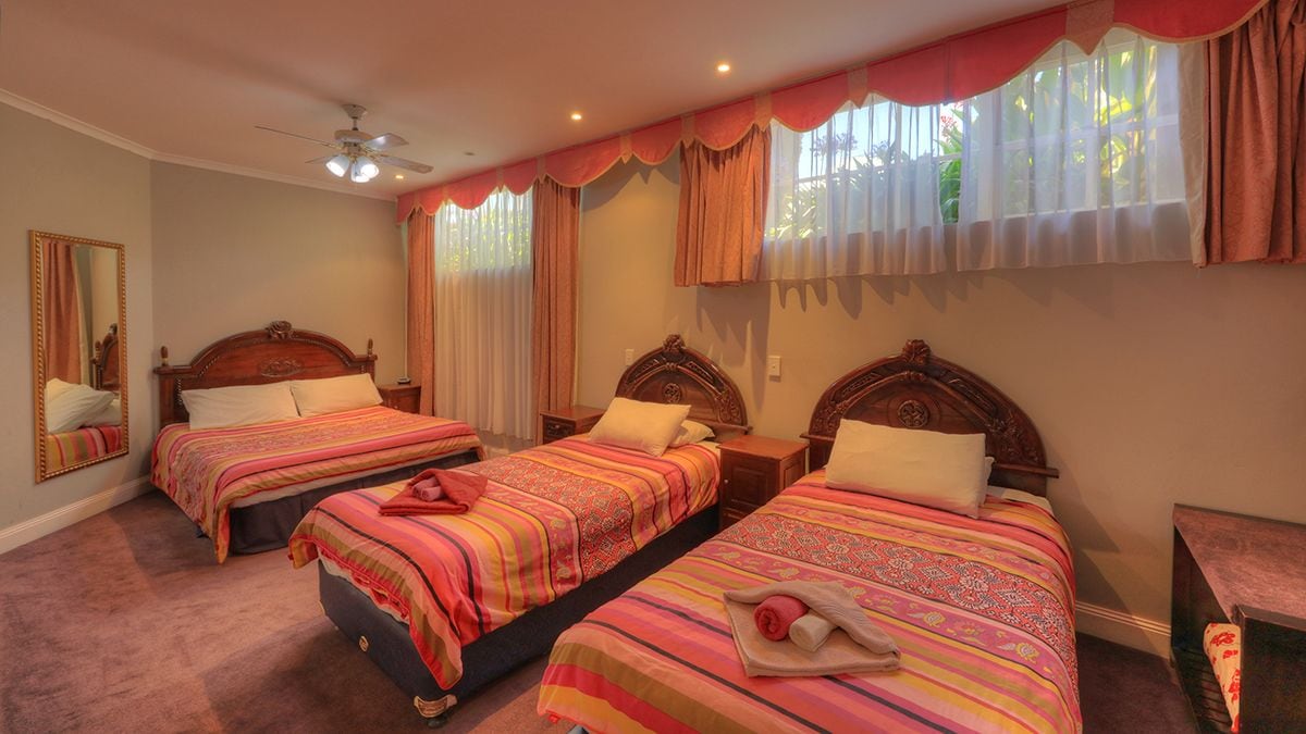Habitación de hotel con tres camas individuales, cada una con cabecera de madera y colcha a rayas. Se observa un ventilador de techo, un espejo grande en la pared izquierda y una ventana con cortinas.