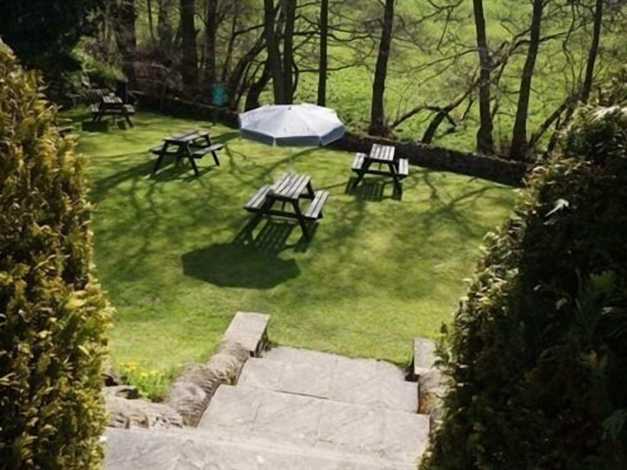 A patio area with stone steps leading up to a grassy lawn, surrounded by trees and featuring picnic tables and an umbrella.