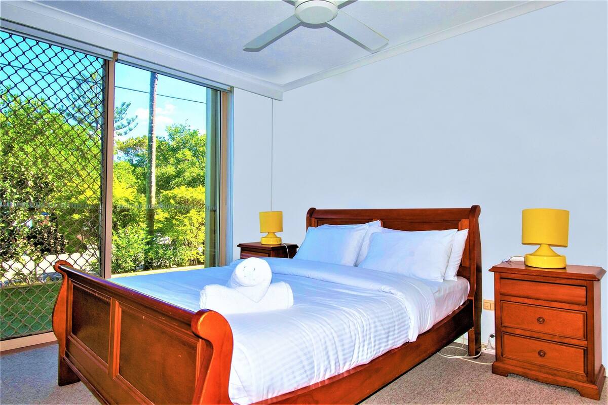 A bedroom with a wooden bed, two bedside tables with lamps, a window with a view of greenery, and a ceiling fan.