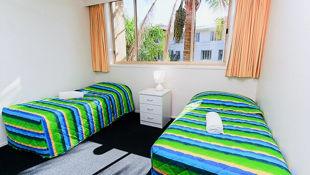 A hotel room with two single beds, a small white dresser, a window with curtains, and a view of palm trees and buildings outside. - Sand Castles on Currumbin Beach (Currumbin)