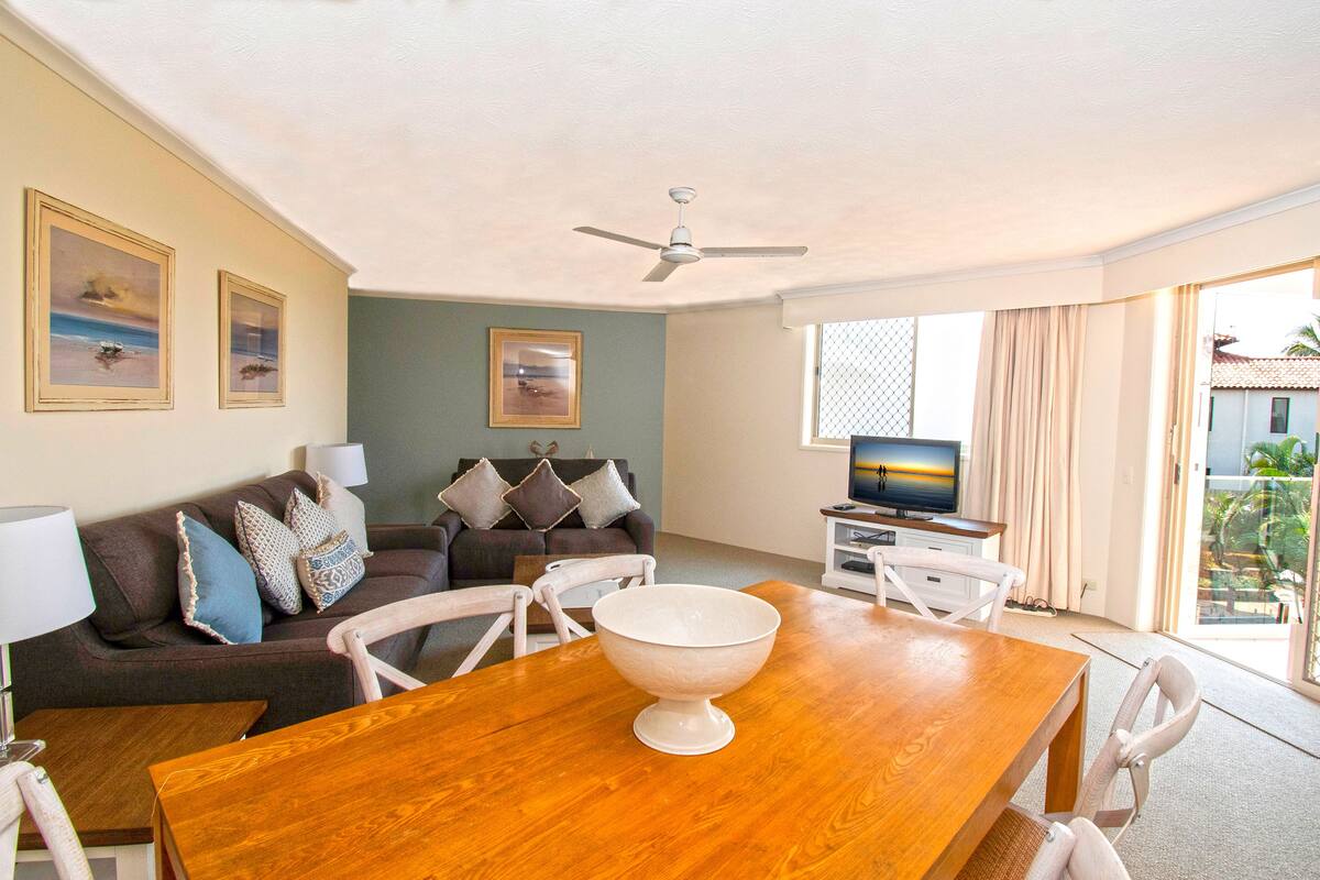 A living room with a wooden dining table, white chairs, a brown sofa with patterned cushions, a ceiling fan, and a television.