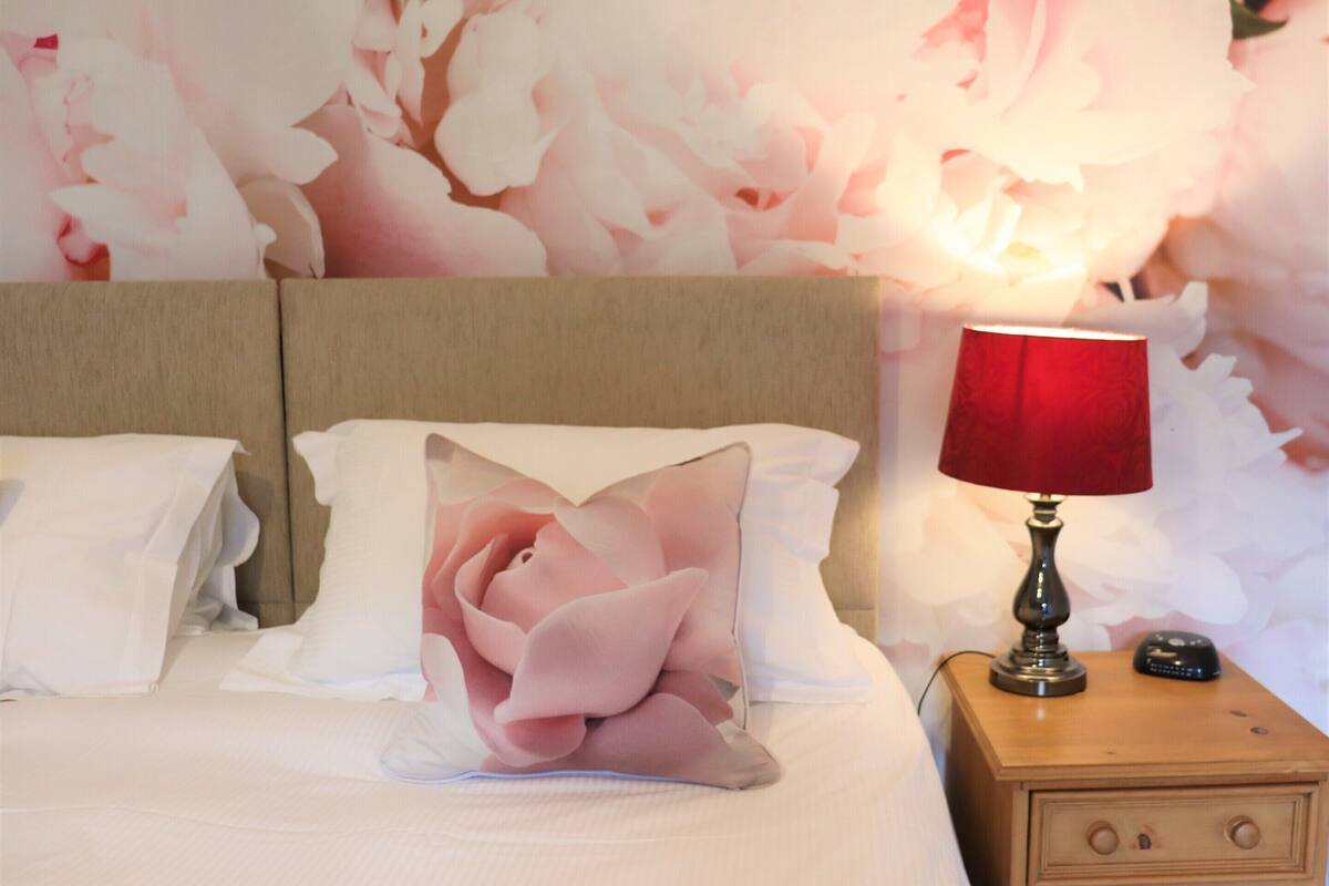 A neatly made bed with white bedding and a pink rose-shaped pillow, a red lamp on a wooden nightstand, and a floral wallpaper background.