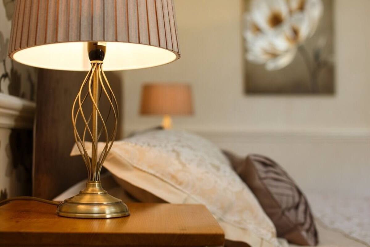 A bedside table with a lamp, a bed with a white and brown blanket, and a painting on the wall.