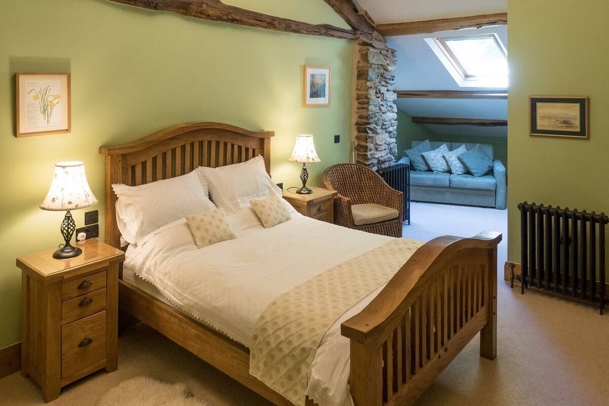 A bedroom with a wooden bed, two bedside tables with lamps, a wicker chair, a stone wall, a radiator, and a view of a living room through an open door.