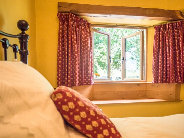 A bedroom with a wooden headboard, a window with curtains, and a view of greenery outside. - The Wayside and Whisky Barn (Millom)