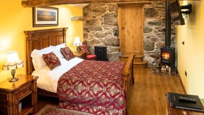 A cozy bedroom with a wooden bed, a stone wall, a fireplace, and a wooden ceiling structure. - The Wayside and Whisky Barn (Millom)