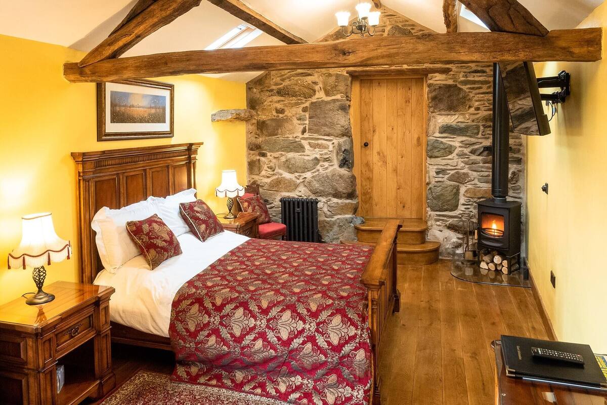 A cozy bedroom with a wooden bed, a stone wall, a fireplace, and a wooden ceiling structure.