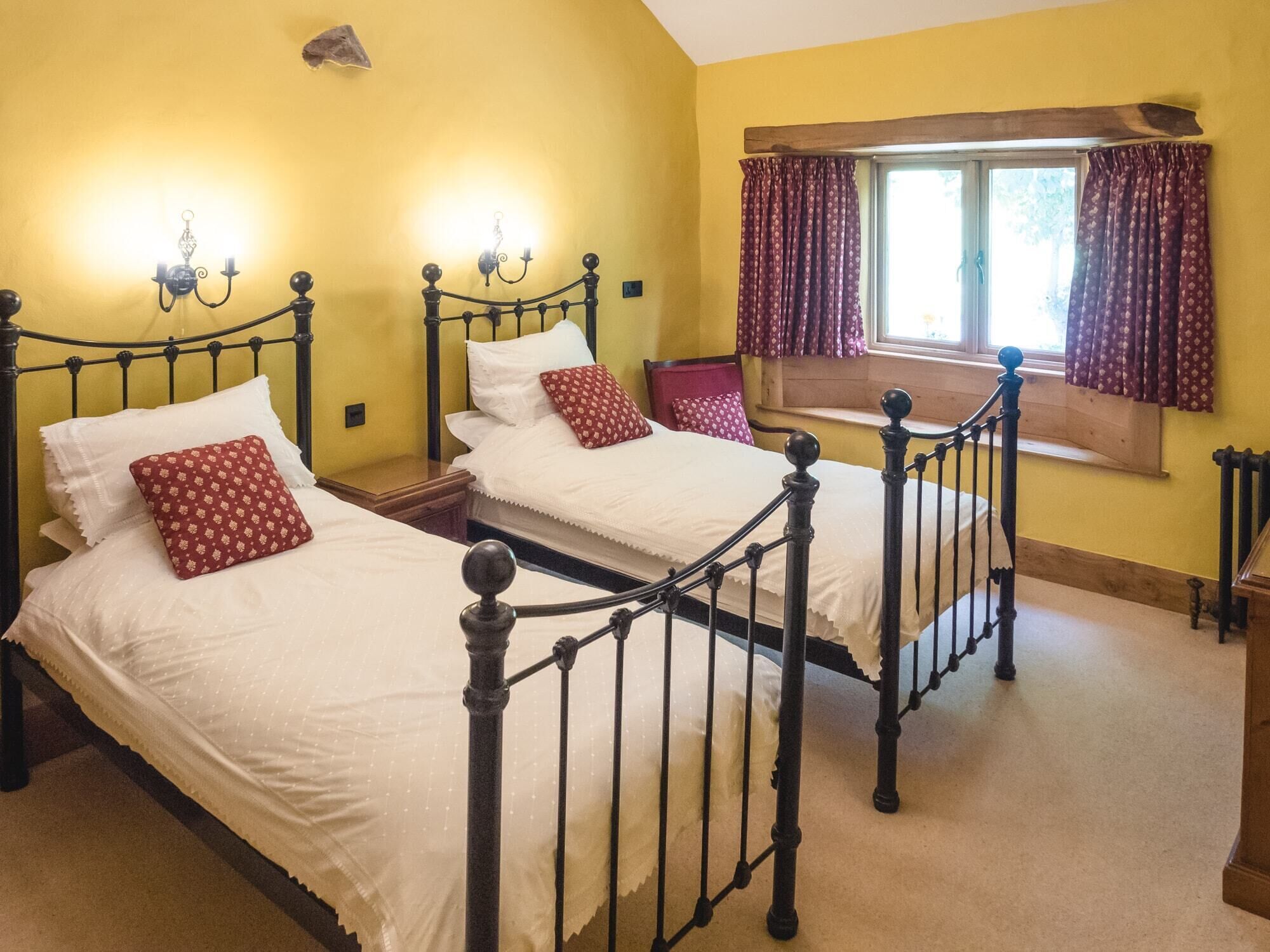 A bedroom with two beds, each with a headboard and a nightstand. There is a window with curtains, and a wall-mounted light fixture above the beds.