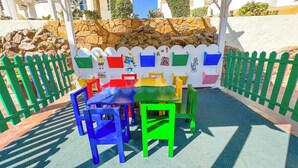 Een kinderspeelplaats met kleurrijke tafels en stoelen, een stenen muur en een wit hekwerk.