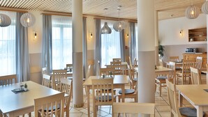 A spacious dining area with wooden tables and chairs, large windows with curtains, and pendant lighting. - JUFA Hotel Pöllau (Poellau)