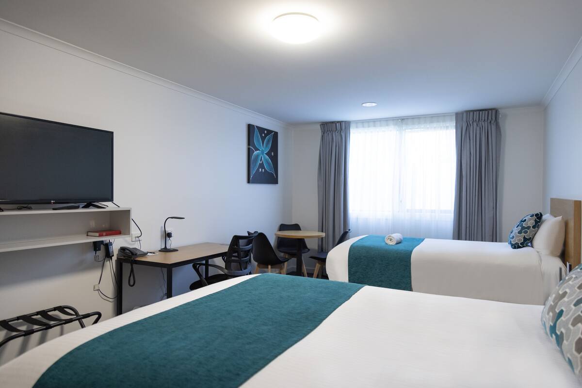 Une chambre d’hôtel avec deux lits, un bureau avec une chaise, une télévision et une fenêtre avec des rideaux.