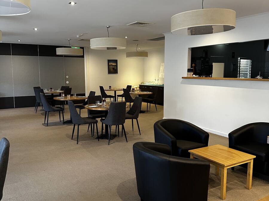 A modern hotel lobby with a dining area, black leather sofas, a wooden table, and a reception desk.