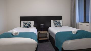A hotel room with two beds, each with a teal blanket and matching pillows, a small bedside table with a lamp, and a window with curtains.