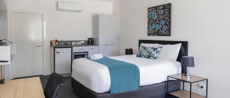 A hotel room with a bed, bedside table, desk, chair, kitchenette, and a painting on the wall.