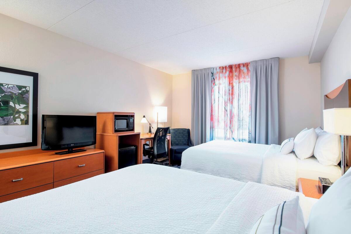 A hotel room with two beds, a desk with a television, a chair, and a window with curtains.