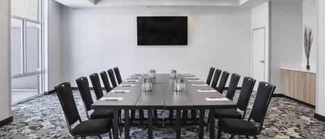 A conference room with a long table, black chairs, a flat-screen TV on the wall, and a patterned carpet.