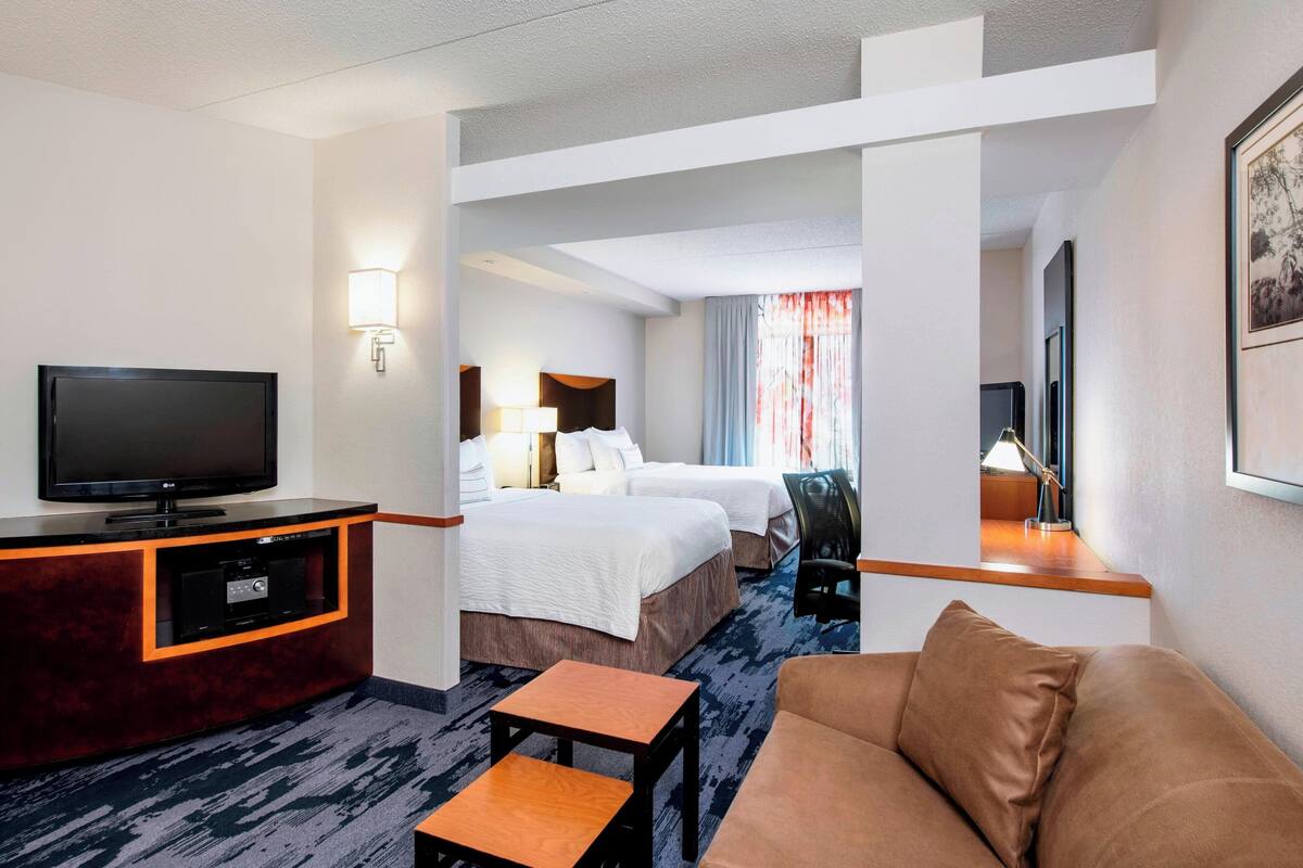 A hotel room with a flat-screen TV, two beds, a sofa, a small table, and a desk with a lamp.