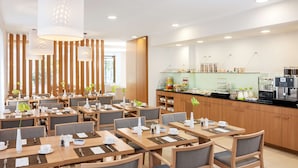 A modern hotel dining area with multiple tables, chairs, and a breakfast bar with various food items and beverages. - NH Frankfurt Messe (Frankfurt)