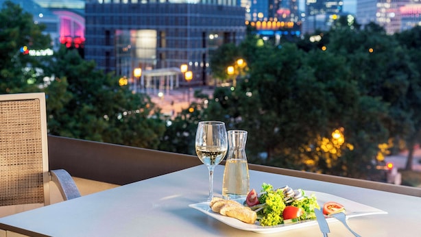 A dining table with a plate of food, a wine glass, and a water bottle, set against a cityscape view. - NH Frankfurt Messe (Frankfurt)