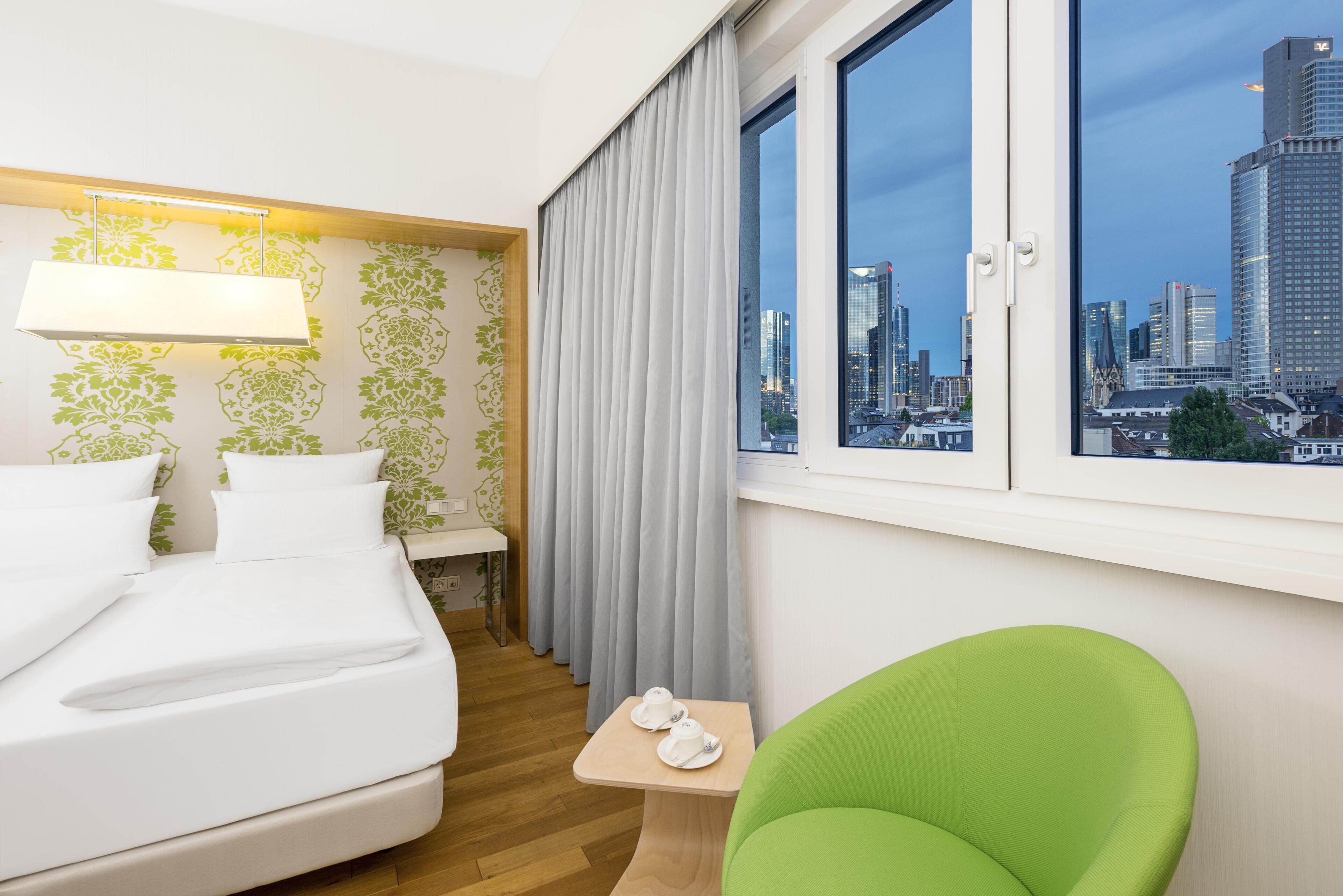 A hotel room with a bed, a green chair, a small table with two white cups, a window with a city view, and a wall with green floral wallpaper.