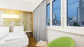 A hotel room with a bed, a green chair, a small table with two white cups, a window with a city view, and a wall with green floral wallpaper.