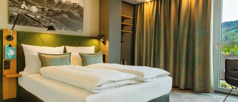 Habitación de hotel moderna con una cama grande, cabecera con panel verde, un cuadro colgado, mesita de noche con lámpara y vista a zonas verdes por la ventana.