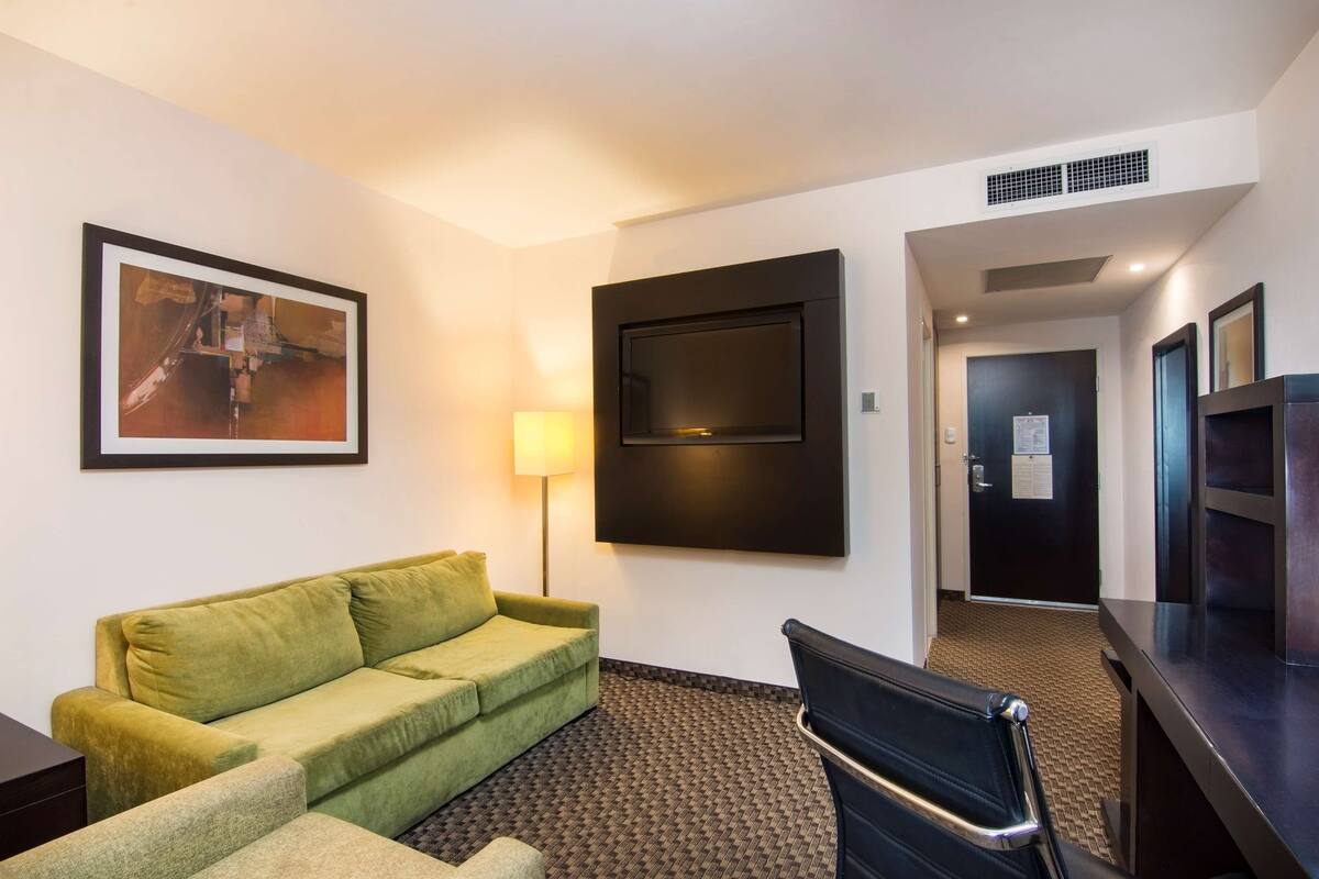Habitación de hotel con sofá verde, una silla negra, televisor de pantalla plana, un cuadro abstracto enmarcado y una lámpara.
