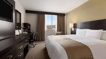 A hotel room with a large bed, a desk with a flat-screen TV, a chair, and a view of the city through the window.