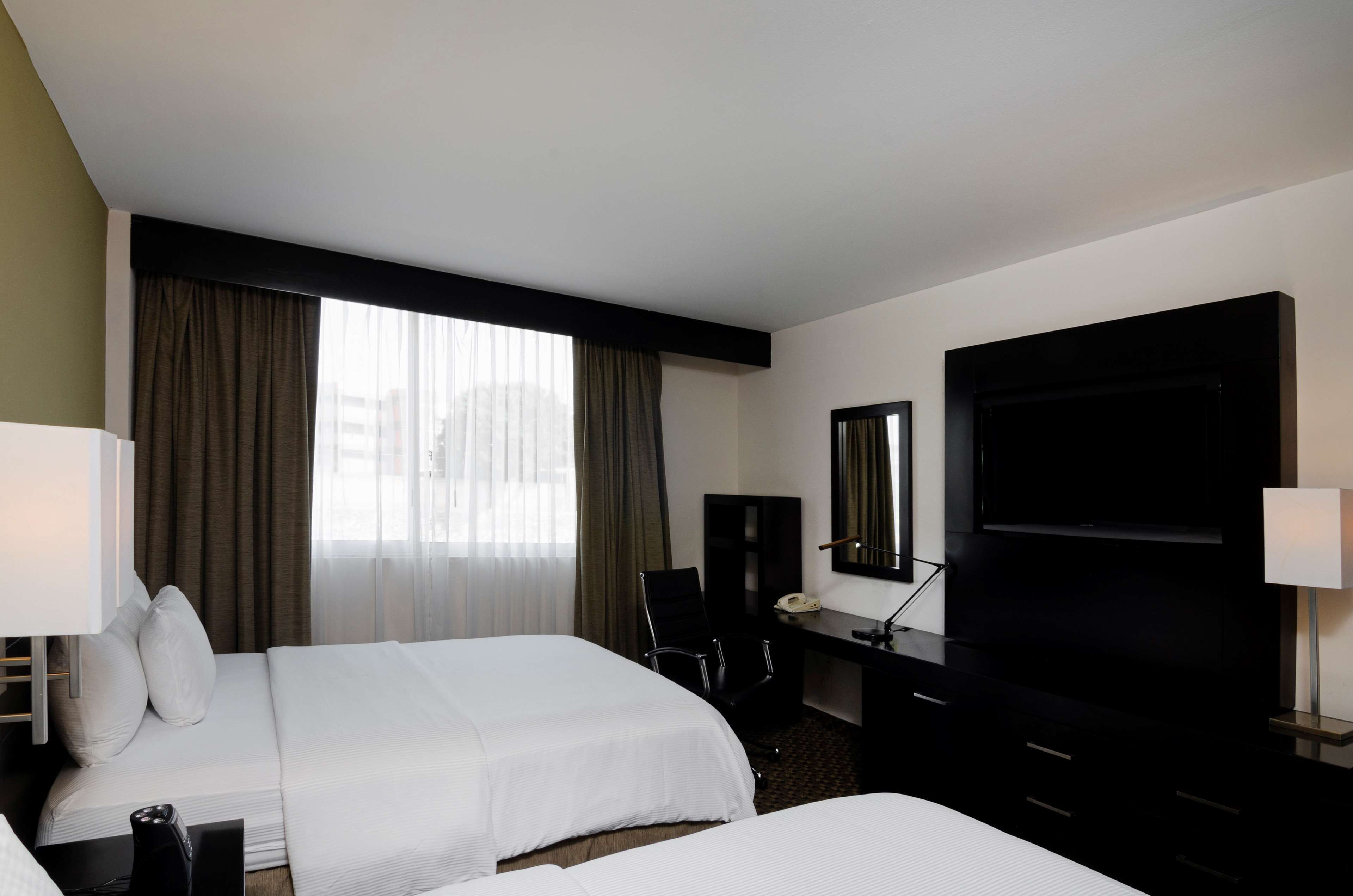 Una habitación de hotel con dos camas, un escritorio con silla, televisión y ventana con cortinas.