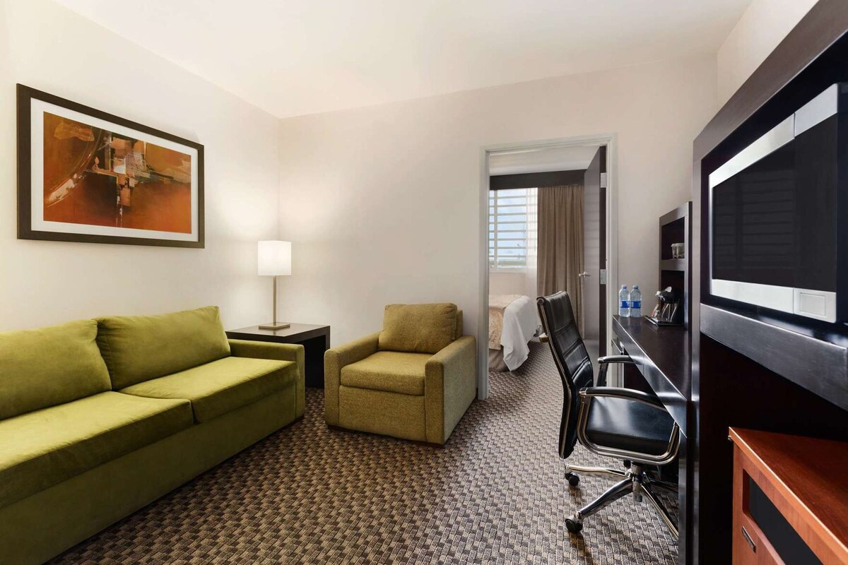 A hotel room with a green sofa, a small armchair, a desk with a chair, a TV, and a painting on the wall.