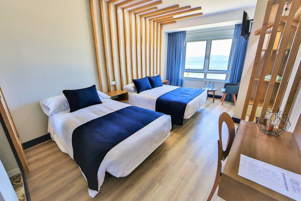 Ein Hotelzimmer mit zwei Betten, Holzpaneelen als Kopfteil, einem kleinen Tisch mit einem Stuhl und einem Fenster mit Blick aufs Meer.