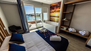 Habitación de hotel con cama, zona de estar con una silla, una pequeña mesa con una botella, un banco con toallas y un ventanal con vistas al mar.