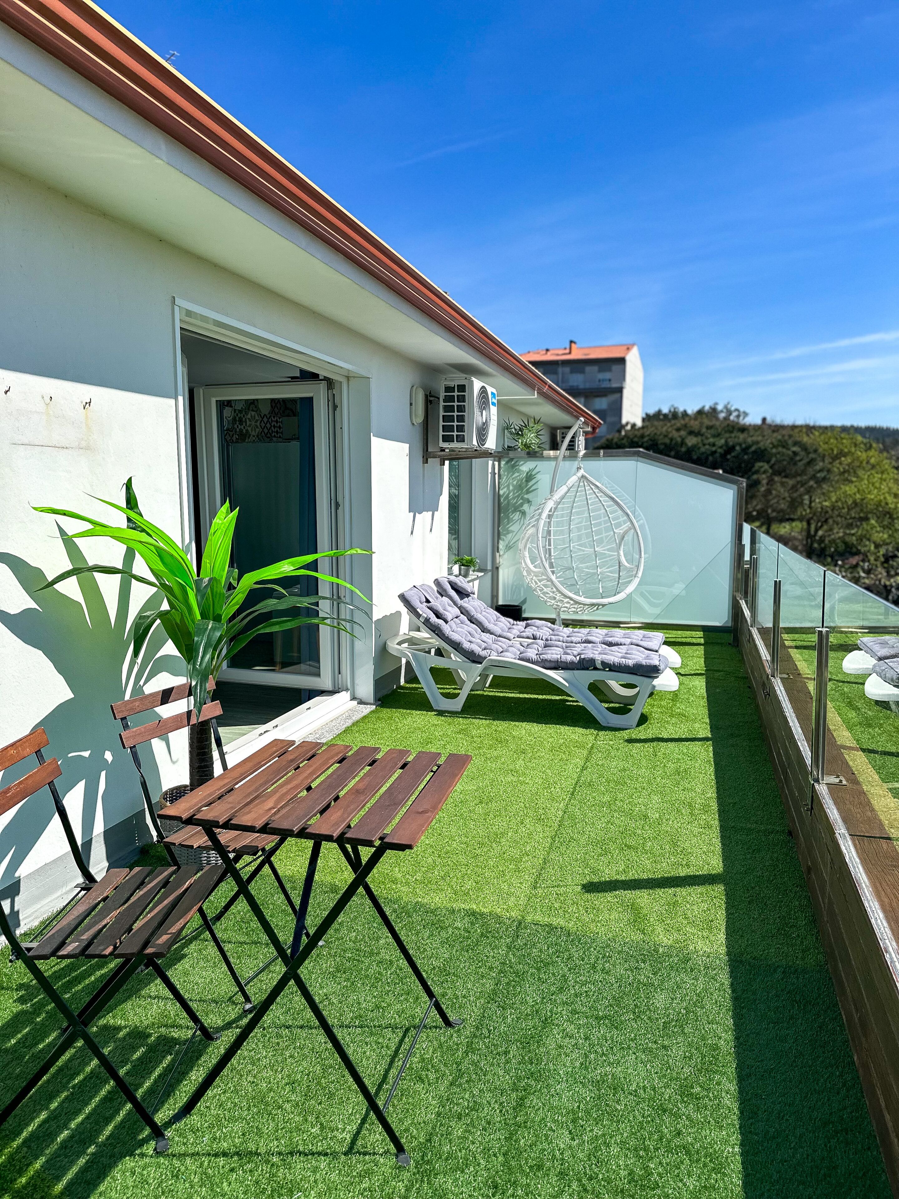 Een balkon met kunstgras, een houten tafel, twee loungestoelen en een airconditioning.