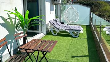 Een balkon met kunstgras, een houten tafel, twee loungestoelen en een airconditioning.