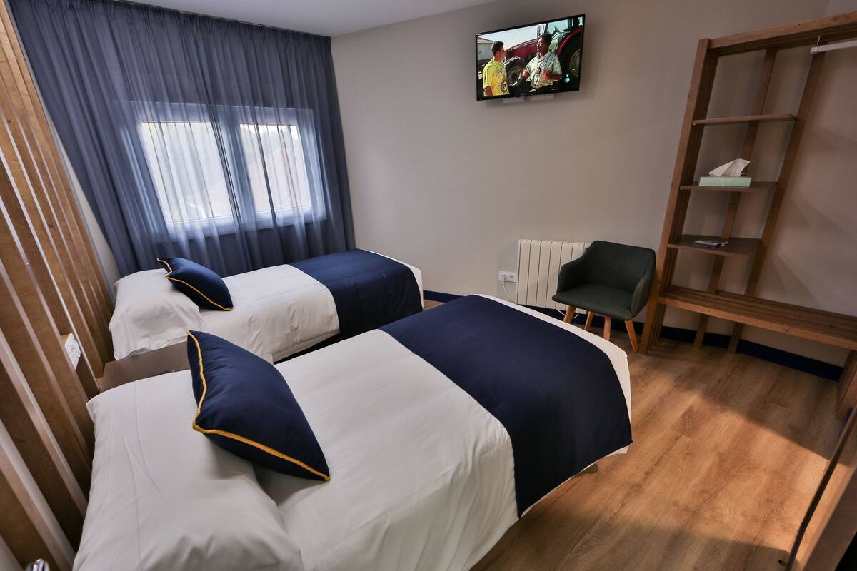 Ein Hotelzimmer mit zwei Betten, einem Wandfernseher, einem Holzregal und einem Fenster mit Vorhängen.