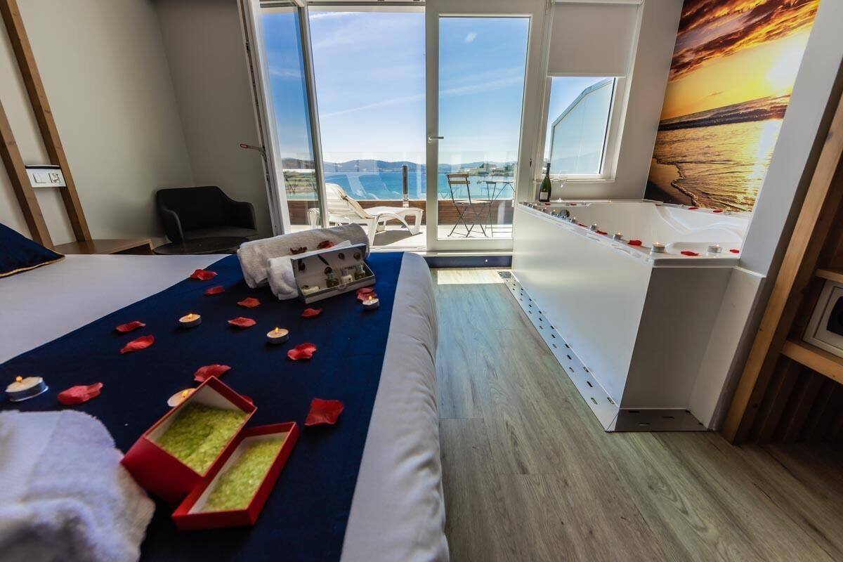 Ein Hotelzimmer mit Bett, einem kleinen Tisch mit Flasche und Kerzen, Meerblick durch große Fenster und einem Gemälde von einem Sonnenuntergang.