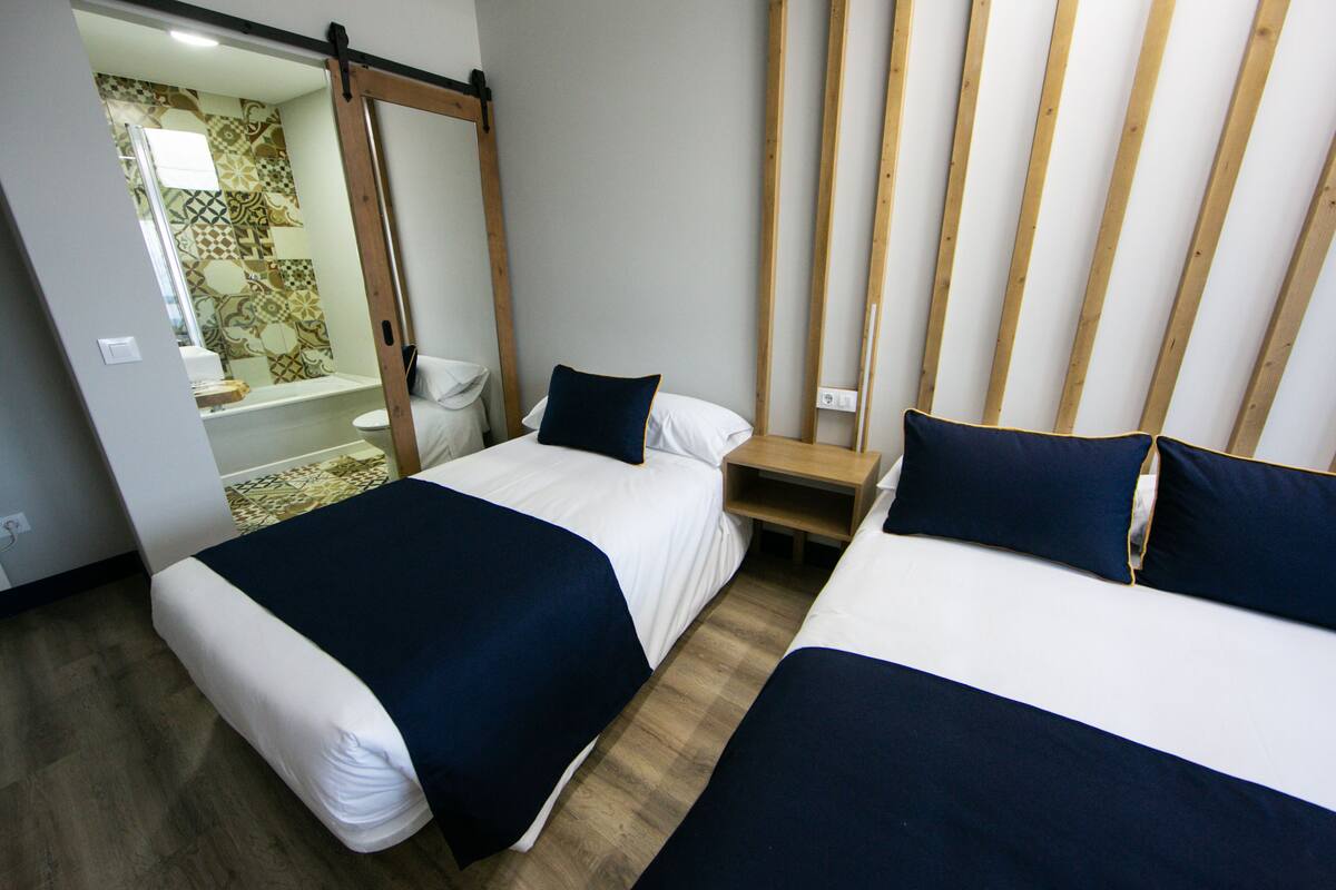 Ein Hotelzimmer mit zwei Betten, einem Holzkopfteil, einem kleinen Nachttisch und einem Badezimmer mit gemusterten Fliesen.