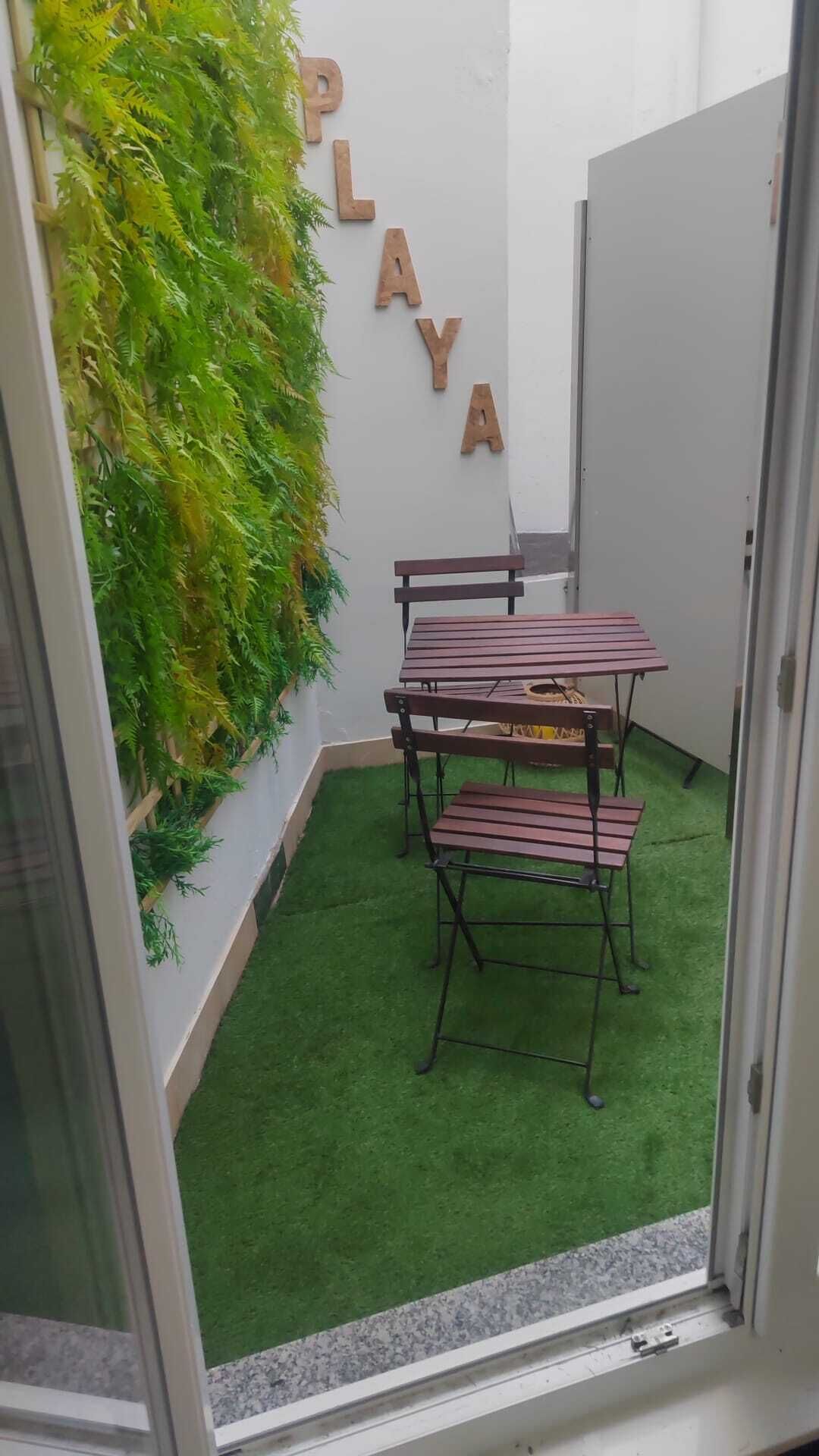 A small patio area with a table and chairs, a green artificial grass floor, and a wall with wooden letters spelling "PLAYA".