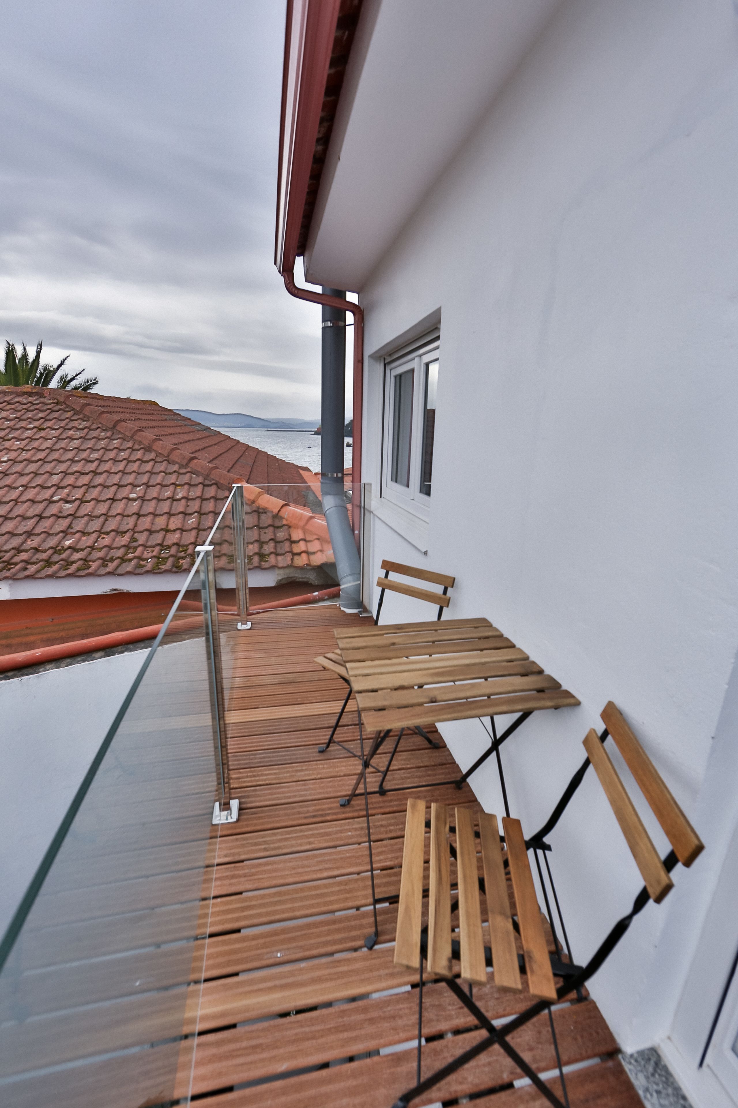 A balcony with a wooden table and chairs, a glass railing, and a view of the sea and a red-tiled roof.