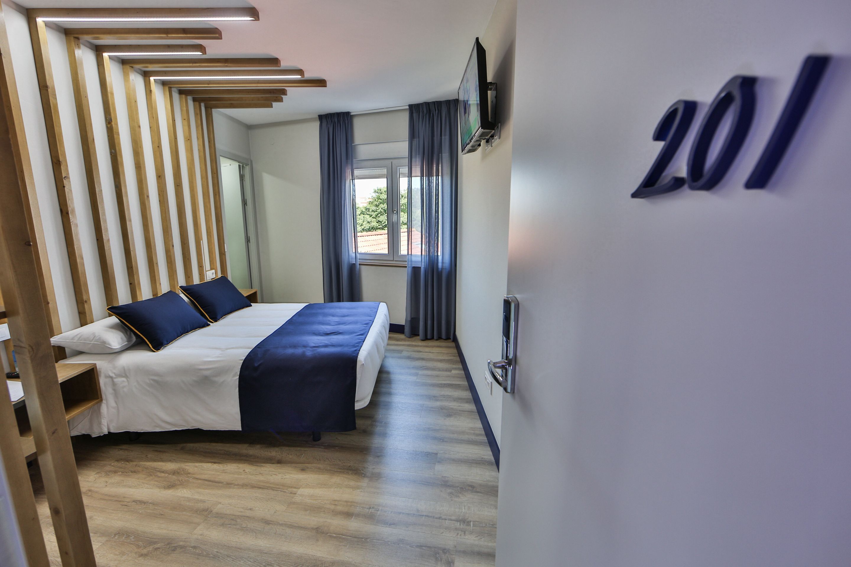 Una habitación de hotel moderna con suelo de madera, una cama con colcha azul y un televisor montado en la pared.