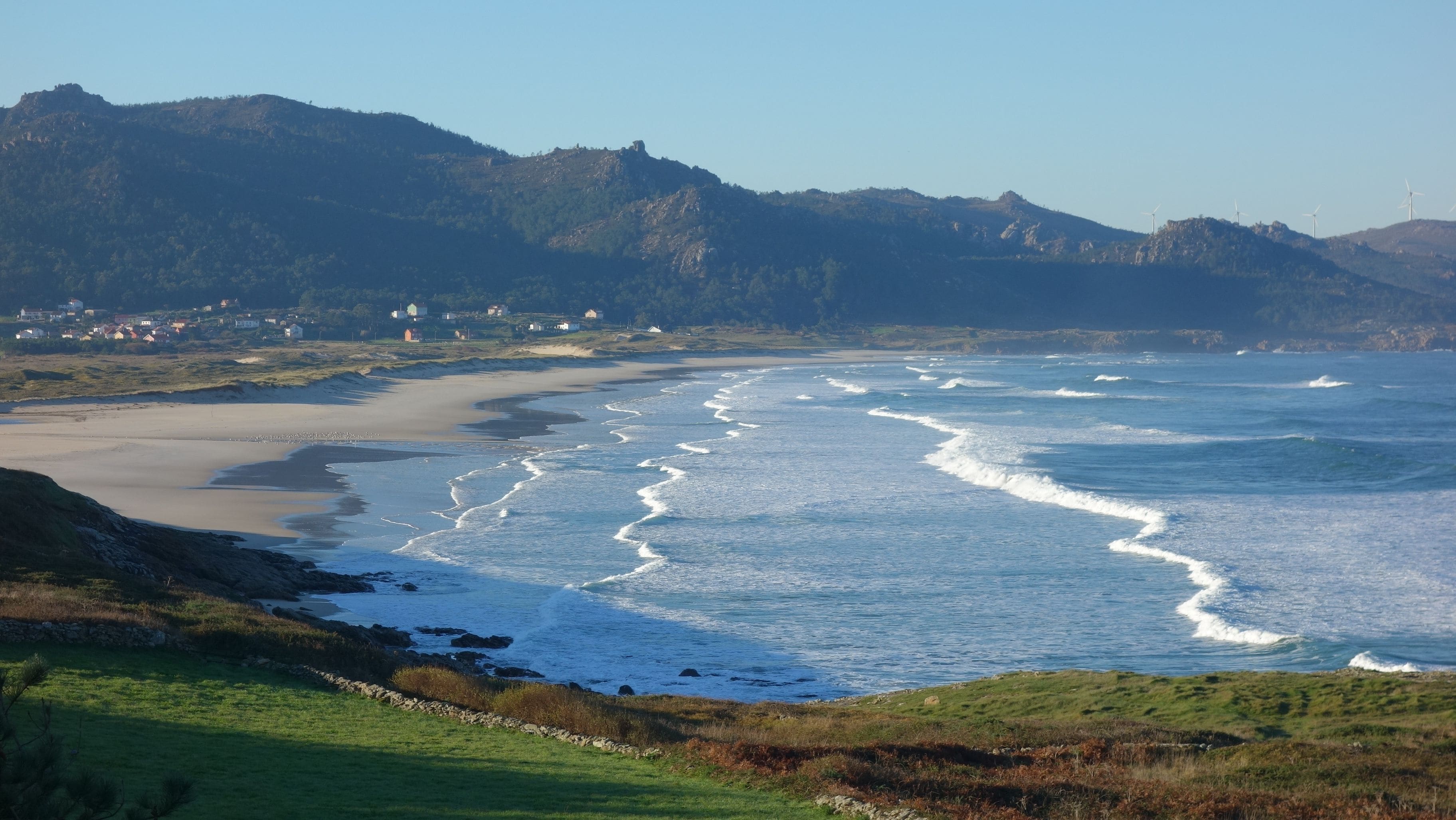 Un paisaje costero con una playa, olas y un pueblo lejano.