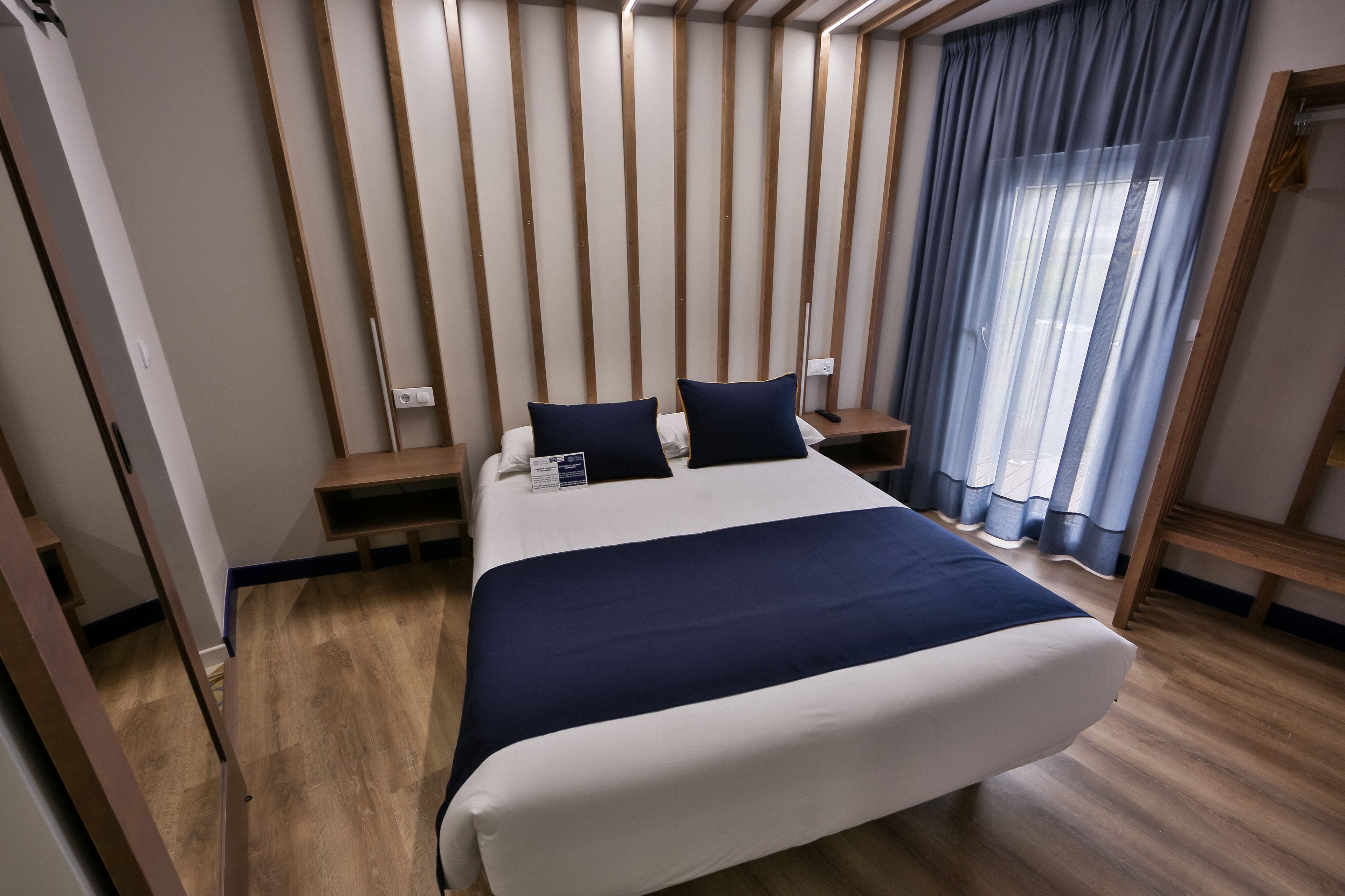 A modern hotel room with a large bed, wooden panel walls, a bedside table, and a window with curtains.